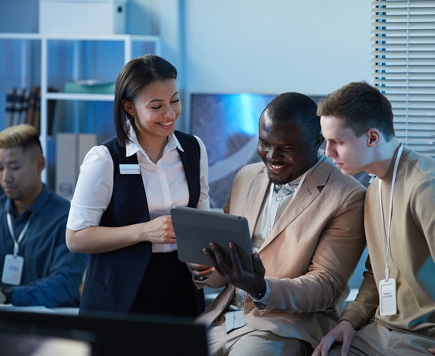 Three people wearing name badges look at a tablet together and smile in an office setting, while another person works in the background.