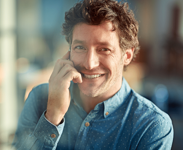 A man with curly hair wearing a blue shirt smiles while talking on a smartphone indoors.