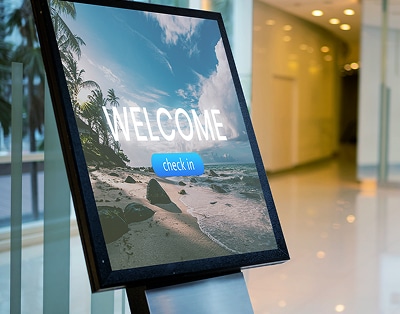 Digital sign in a modern lobby displays a beach scene with the words "WELCOME" and a blue "check in" button.
