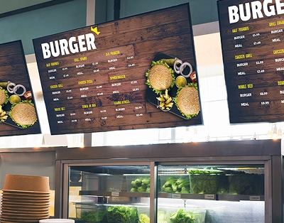 Digital menu boards above a counter display various burger options and prices at a restaurant, with a salad bar visible below.