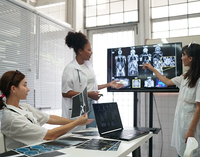 Three medical professionals analyze and discuss body scan images displayed on a large monitor in a clinical setting.