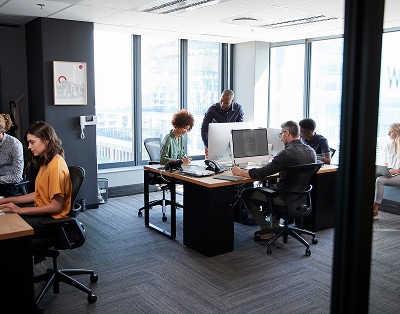 Several people work at desks with computers in a modern office with large windows and city views during the daytime.