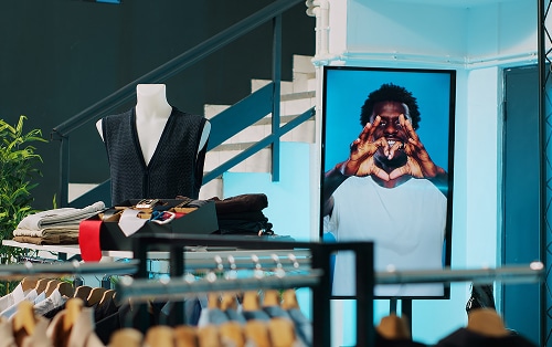 A clothing store display with folded clothes, a vest on a mannequin, and a digital screen showing a person making a heart shape with their hands.