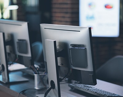 Two computer monitors are placed side by side on a desk in an office setting, with blurred office equipment and a screen visible in the background.