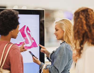Three women stand in front of a digital map display, discussing directions while one holds a smartphone.