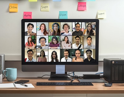 A computer monitor displays a video conference with 16 participants. The desk holds a keyboard, mouse, mug, phone, and sticky notes are on the wall behind the screen.