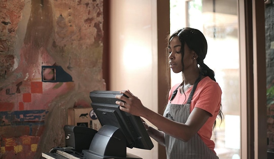 A woman wearing an apron stands at a point-of-sale terminal in a rustic cafe, operating the touchscreen register.