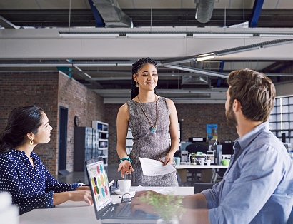 Three coworkers have a discussion at a desk in a modern office. One woman stands holding papers, while the other two are seated, one using a laptop.
