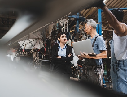 A woman in business attire stands with a man holding a laptop and another worker in overalls inside an industrial workshop.