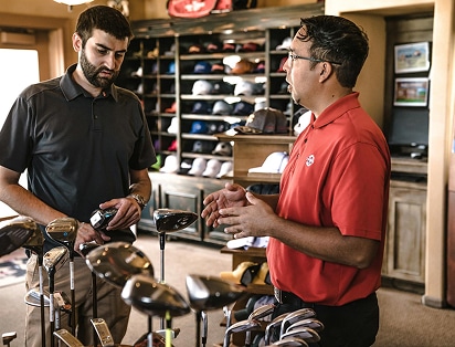 Two men stand in a golf pro shop discussing golf clubs, with shelves of hats and golf gear in the background.