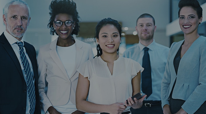 Five professionally dressed people stand together in an office, facing the camera and smiling.