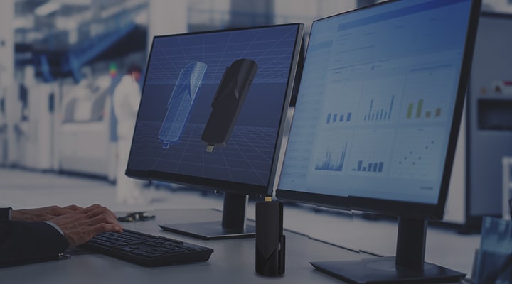 A person works at a desk with two computer monitors displaying a 3D model and data charts in a modern office or lab setting.