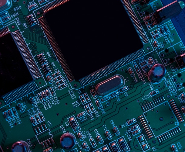 Close-up view of a green circuit board with various electronic components, including microchips, resistors, and capacitors, illuminated by blue and pink lighting.