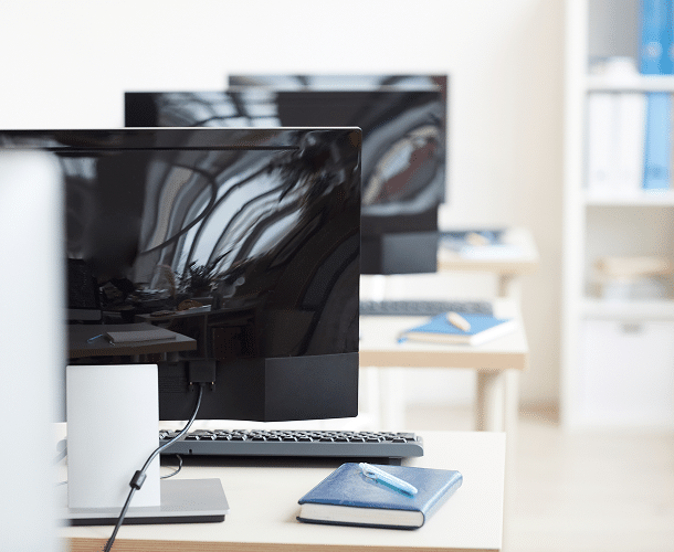Row of desktop computers on desks in a bright office or classroom, with notebooks and pens placed on the desks.