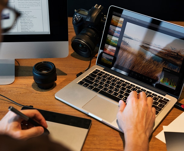 Person editing a landscape photo on a laptop using photo editing software, with a graphics tablet, camera, and lens on a wooden desk.