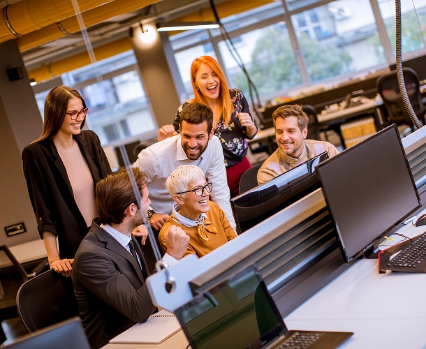 Six people in business attire gather around a computer workstation in a modern office, smiling and looking at the screen together.