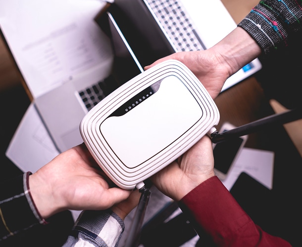 Three people holding a white wireless router together above a desk with laptops and scattered papers.