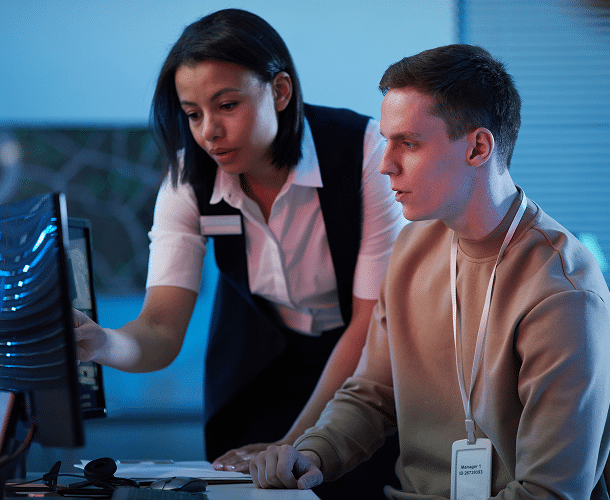 Two people in an office setting look at a computer monitor; one is pointing at the screen while the other sits and listens attentively.