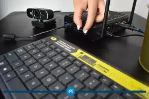 A hand plugs a cable into a black electronic device on a desk, with a keyboard and a webcam nearby.