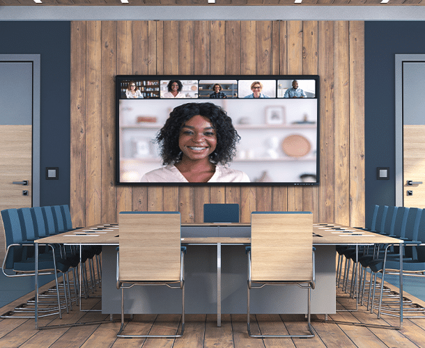 A conference room with a large screen displaying a video call; one woman is shown prominently, with five other participants in smaller windows above.