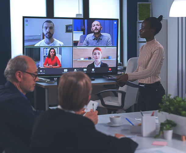 A group of people in an office have a video conference with four colleagues displayed on a large screen. A woman stands presenting, while two others are seated at the table.