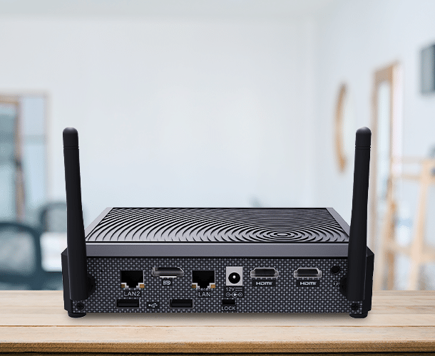 A black mini PC with two antennas sits on a wooden surface, showing ports for LAN, HDMI, power, and USB; a blurred office environment is in the background.