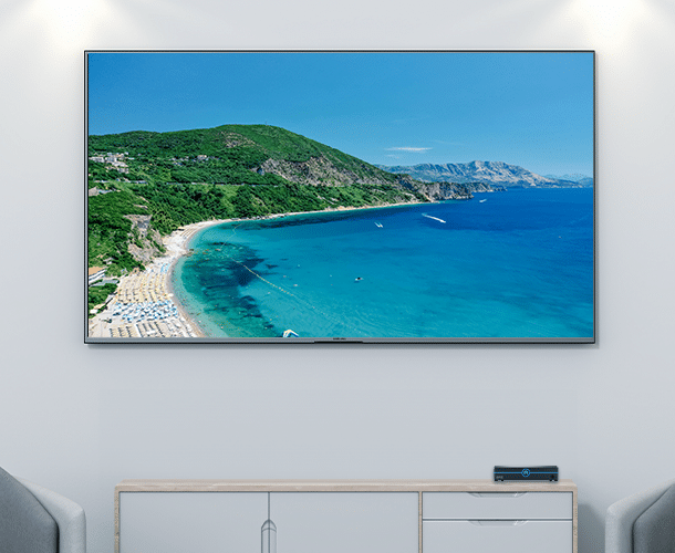 A large wall-mounted TV displays a scenic coastal landscape with a turquoise sea, sandy beach, and green hills; modern living room furniture is visible below the screen.