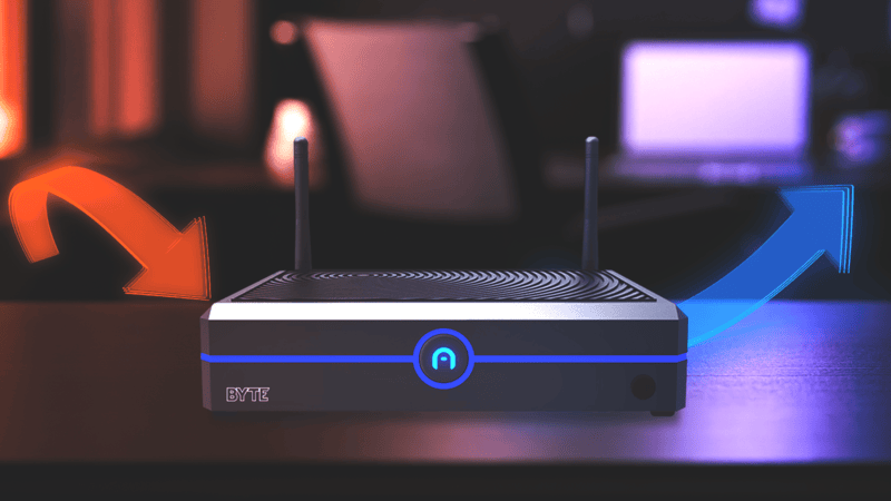 A wireless router with two antennas emits an orange signal to the left and a blue signal to the right, placed on a desk with blurred office equipment in the background.