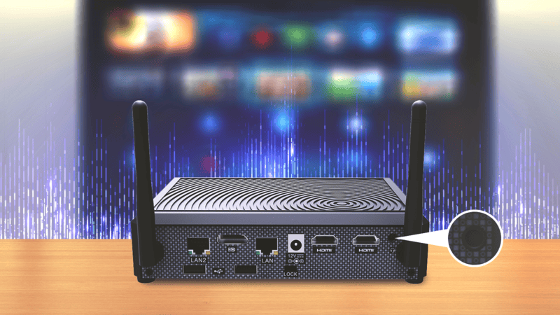 A small black electronic device with two antennas sits on a wooden surface, showing various ports on the back, with a blurred screen in the background.