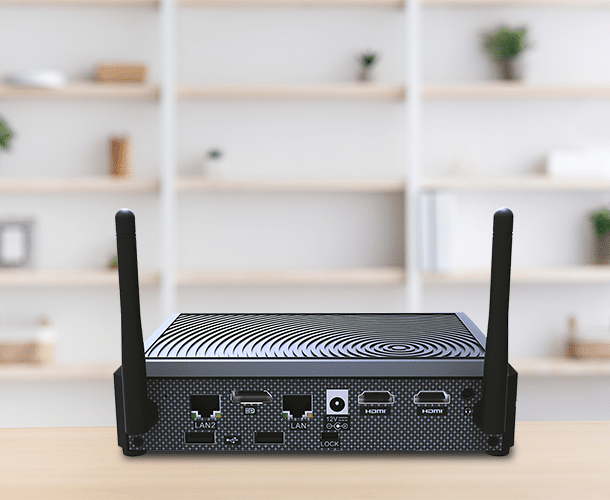 A compact black network device with two antennas and multiple ports sits on a wooden desk with blurred shelves in the background.