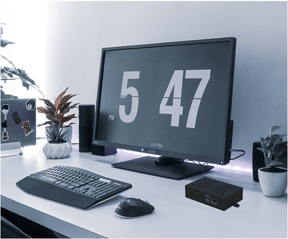 A clean white desk setup with a computer monitor displaying the time 5:47 PM, keyboard, mouse, plants, a mini PC, and a laptop with stickers.