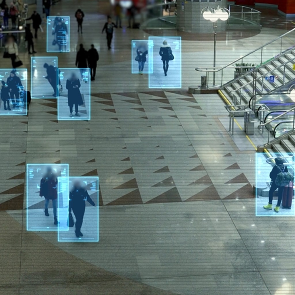 A view of people walking in a large indoor space, with digital blue rectangles highlighting individuals, suggesting surveillance or facial recognition technology.